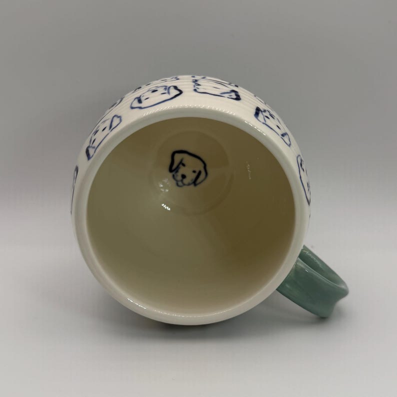 May include: White ceramic mug with a light green handle. The mug is decorated with blue line drawings of dog faces. A single dog face is drawn on the inside bottom of the mug.