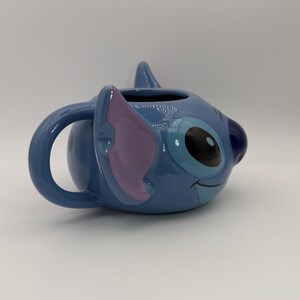 Puede incluir: Una taza de cer&aacute;mica azul con la forma del personaje Stitch. La taza presenta la cara de Stitch, con grandes ojos negros, una oreja morada y una expresi&oacute;n sonriente. El asa tiene forma curva azul.