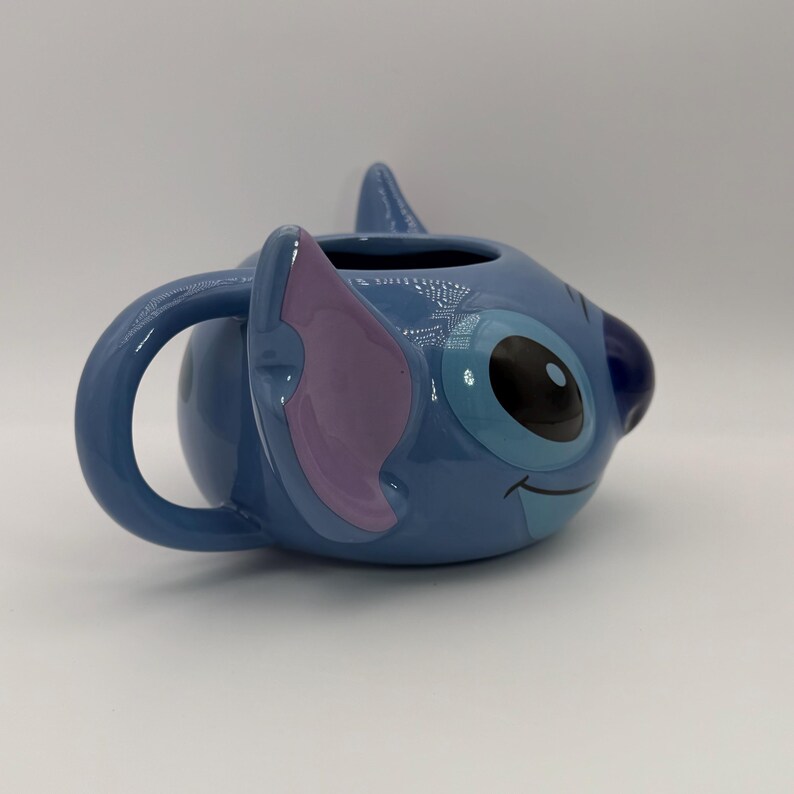 Puede incluir: Una taza de cer&aacute;mica azul con la forma del personaje Stitch. La taza presenta la cara de Stitch, con grandes ojos negros, una oreja morada y una expresi&oacute;n sonriente. El asa tiene forma curva azul.