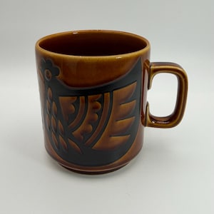 May include: A brown ceramic mug featuring a dark brown embossed graphic of a stylised bird on the side. The mug has a glossy finish, a cylindrical body, and a sturdy handle on the right side. The interior and exterior display a warm, earthy tone.
