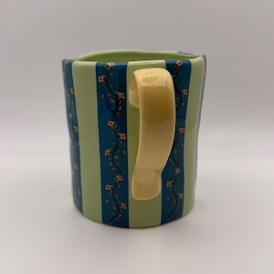 May include: A ceramic mug with a cream-colored handle. The mug features alternating vertical stripes of light green and dark blue. Floral vines with small red flowers and green leaves decorate the blue stripes. The mug is a unique and decorative item.