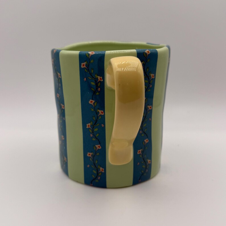 May include: A ceramic mug with a cream-colored handle. The mug features alternating vertical stripes of light green and dark blue. Floral vines with small red flowers and green leaves decorate the blue stripes. The mug is a unique and decorative item.
