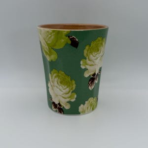 Puede incluir: Un vaso verde con estampado floral. El vaso presenta un patr&oacute;n repetido de rosas blancas y verde claro sobre un fondo verde oscuro. El borde del vaso es de color marr&oacute;n claro.