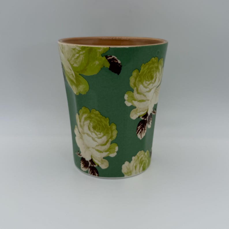 Puede incluir: Un vaso verde con estampado floral. El vaso presenta un patr&oacute;n repetido de rosas blancas y verde claro sobre un fondo verde oscuro. El borde del vaso es de color marr&oacute;n claro.