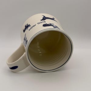 K&ouml;nnte beinhalten: Cremefarbene Keramiktasse mit blauem Rand und Henkel. Die Tasse zeigt blaue Hundeillustrationen und die Worte "DOG MOM" in Blau. Das Innere der Tasse ist hellbeige.