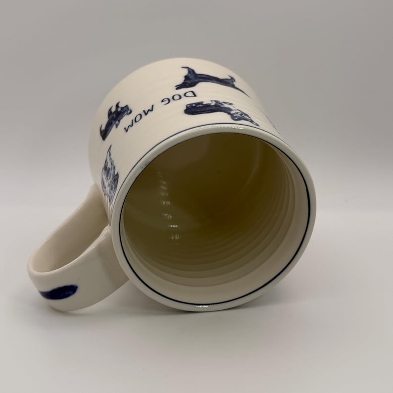 K&ouml;nnte beinhalten: Cremefarbene Keramiktasse mit blauem Rand und Henkel. Die Tasse zeigt blaue Hundeillustrationen und die Worte "DOG MOM" in Blau. Das Innere der Tasse ist hellbeige.