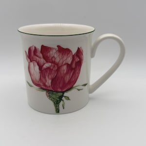 May include: A white ceramic mug featuring a detailed illustration of a large, deep red rose with green leaves and a stem. The mug has a smooth, cylindrical body, a curved handle on the right side, and a thin dark green rim around the top edge.