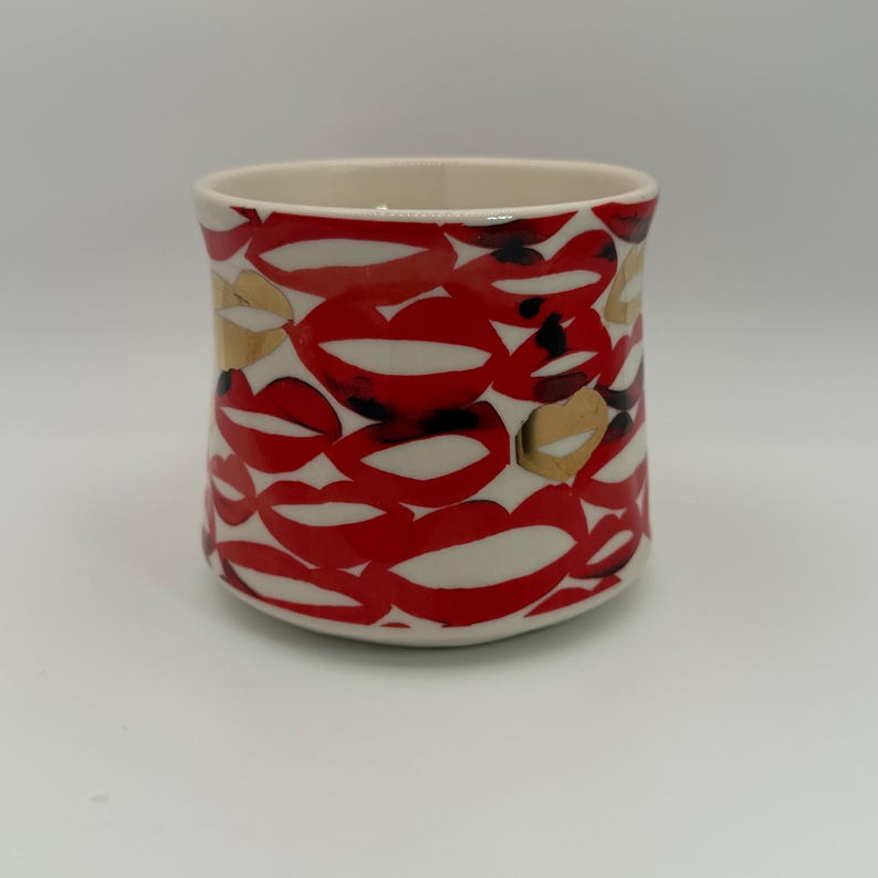 Pode incluir: Uma caneca cil&iacute;ndrica de cer&acirc;mica com interior e exterior brancos cobertos por um padr&atilde;o de l&aacute;bios vermelhos com realces brancos e detalhes dourados. A caneca est&aacute; sobre um fundo branco.