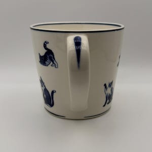 May include: White ceramic mug with a navy blue rim and handle. The mug features navy blue cat illustrations in various poses. The mug is a standard size for coffee or tea.