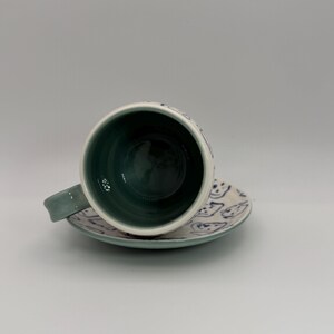May include: A ceramic teacup and saucer set. The cup and saucer are white with blue painted designs. The interior of the cup is a deep teal color, and the handle is a matching teal. The saucer has a matching teal rim.