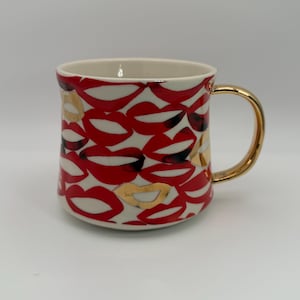 Pode incluir: Uma caneca de cer&acirc;mica com fundo branco e um padr&atilde;o de l&aacute;bios vermelhos. Os l&aacute;bios t&ecirc;m sombreamento preto e detalhes dourados. A al&ccedil;a &eacute; dourada. O interior da caneca &eacute; creme.