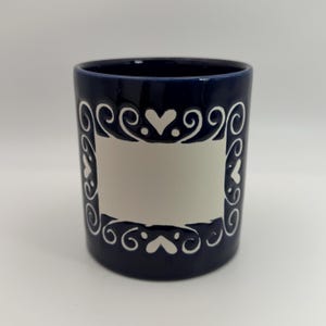 May include: A dark blue ceramic mug featuring a prominent white rectangular center panel. The panel is framed by a decorative white border consisting of swirling scrollwork patterns and small heart shapes, creating a distinct contrast against the deep blue background.