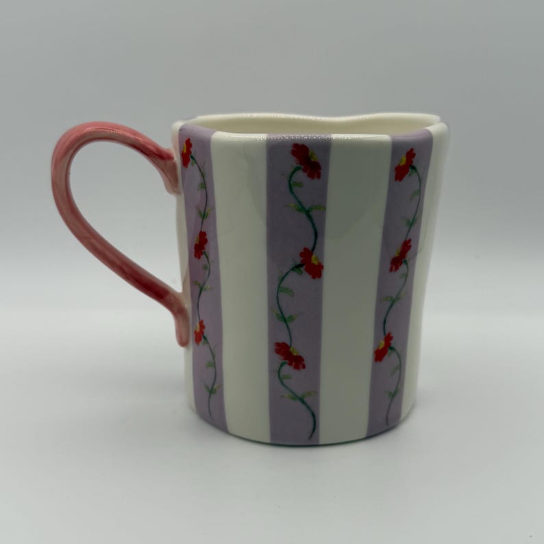 Pu&ograve; includere: Tazza in ceramica con un motivo a righe bianche e lavanda. Fiori rossi con viti verdi sono dipinti tra le strisce. Il manico &egrave; di colore rosa pieno.