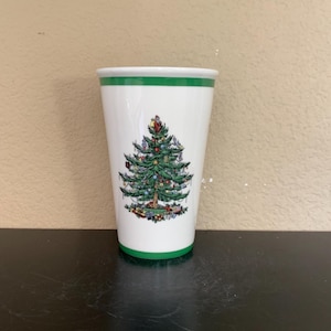 以下が含まれることがあります： 緑色の縁と底が付いた白いセラミックタンブラー。タンブラーには、装飾されたクリスマスツリーの詳細なイラストが描かれています。オーナメント、ギフト、星が木を飾り、お祝いのホリデーデザインを作成します。