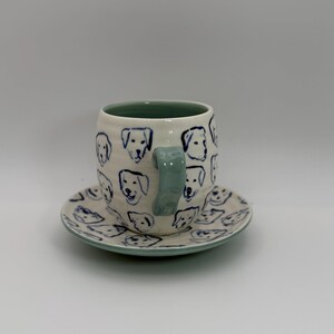 May include: A ceramic coffee cup and saucer set. The cup and saucer are white with blue dog face illustrations. The cup handle and the rim of the cup and saucer are light blue. The cup is cylindrical.