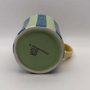 May include: A ceramic mug with a light green interior and a pale yellow handle. The exterior features alternating vertical stripes of light green and blue, with a floral pattern on the blue stripes. The bottom of the mug has the text "by ANTHROPOLOGIE".