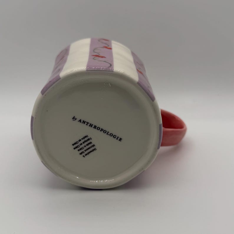Pu&ograve; includere: Una tazza in ceramica con manico rosa e motivo a righe bianche e lavanda. La tazza ha la scritta "by ANTHROPOLOGIE" sul fondo. La tazza &egrave; lavabile in lavastoviglie e adatta al microonde.