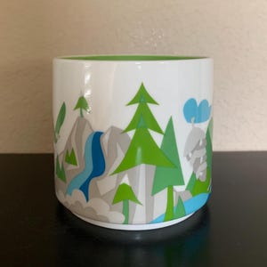 May include: White ceramic mug with a green interior and a landscape design. The design features green trees, grey mountains, a blue waterfall, and blue clouds. The mug is cylindrical and sits on a black surface.