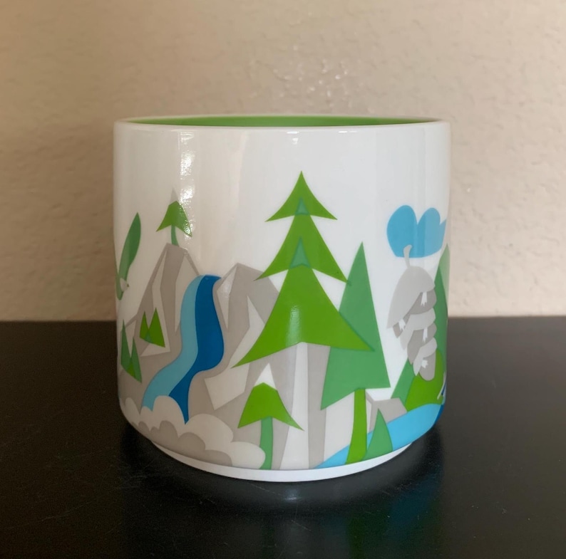 May include: White ceramic mug with a green interior and a landscape design. The design features green trees, grey mountains, a blue waterfall, and blue clouds. The mug is cylindrical and sits on a black surface.