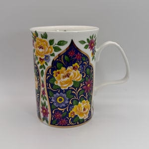 May include: White ceramic mug with a floral design. The mug features yellow, pink, and purple flowers on a dark blue background with gold accents. The handle is white, and the mug has a classic shape.