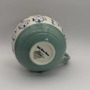 May include: A ceramic mug with a light green and white design. The mug features a light green, ribbed base and a white upper section adorned with blue dog illustrations. The mug has a handle and text that reads "Leah Reena Goren".