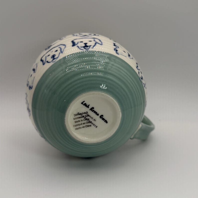 May include: A ceramic mug with a light green and white design. The mug features a light green, ribbed base and a white upper section adorned with blue dog illustrations. The mug has a handle and text that reads "Leah Reena Goren".