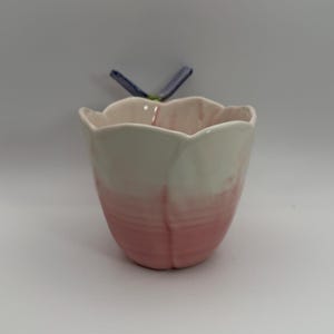 May include: A ceramic tulip-shaped container with a pink and white ombre design. The container has a scalloped rim and a small blue and green accent piece attached to the back.