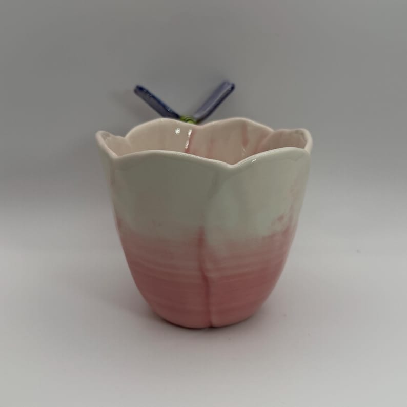 May include: A ceramic tulip-shaped container with a pink and white ombre design. The container has a scalloped rim and a small blue and green accent piece attached to the back.