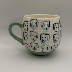 May include: A ceramic mug with a light green handle and base. The mug's off-white surface is decorated with blue line drawings of dog faces. The mug has a rounded shape and a comfortable handle.