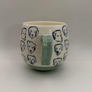 May include: Cream-colored ceramic mug with a light green handle and base. The mug is adorned with blue line drawings of dog faces. The mug has a rounded shape and a textured surface.