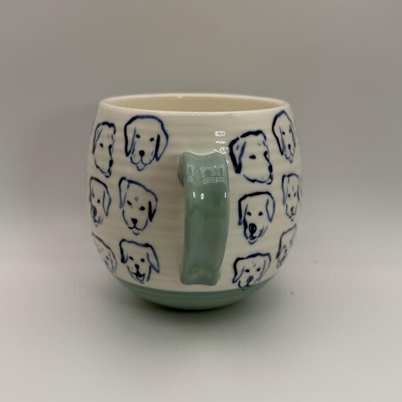 May include: Cream-colored ceramic mug with a light green handle and base. The mug is adorned with blue line drawings of dog faces. The mug has a rounded shape and a textured surface.