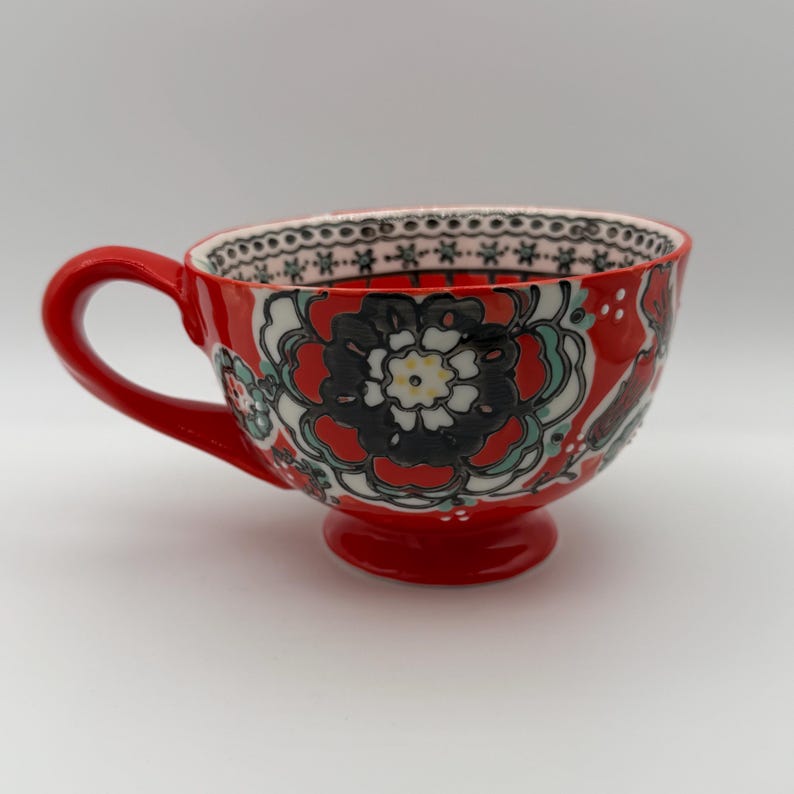 Może przedstawiać: Czerwony ceramiczny kubek do herbaty z kwiatowym wzorem. Kubek ma duży uchwyt i podstawę. Kwiatowy wz&oacute;r zawiera czarne, białe i turkusowe akcenty. Obrzeże kubka ma dekoracyjną czarno-białą obw&oacute;dkę.