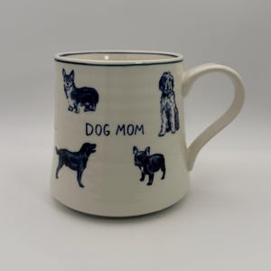 K&ouml;nnte beinhalten: Cremefarbene Keramiktasse mit dunkelblauem Rand und Henkel. Die Tasse zeigt dunkelblaue Illustrationen verschiedener Hunderassen und den Text "DOG MOM".