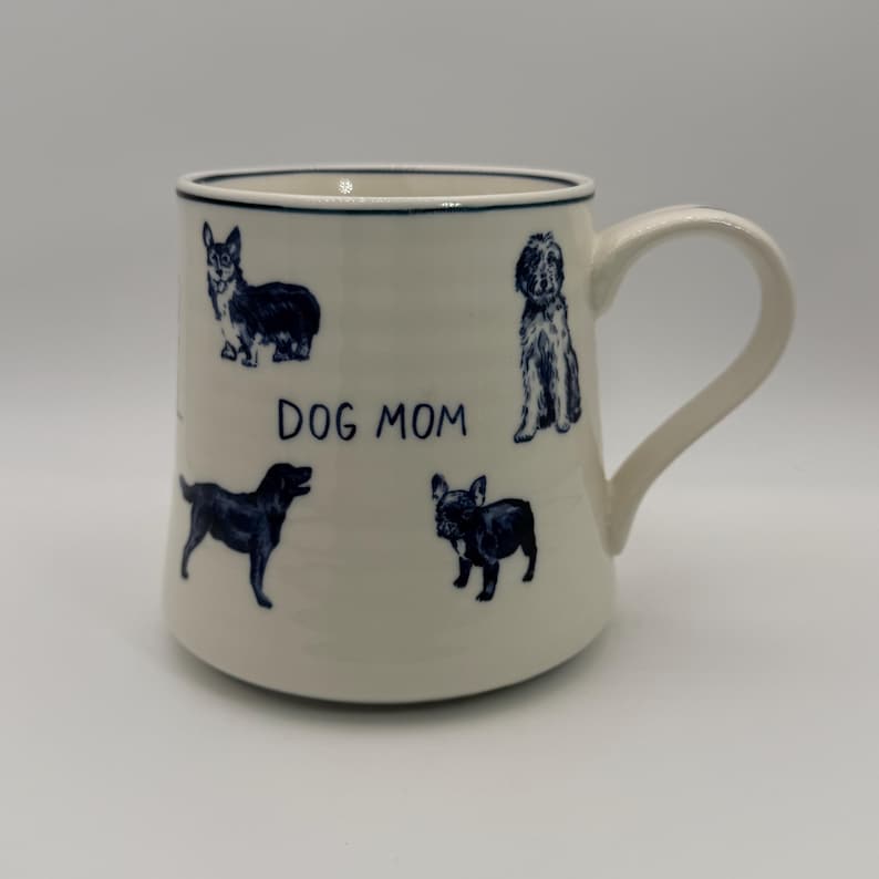 K&ouml;nnte beinhalten: Cremefarbene Keramiktasse mit dunkelblauem Rand und Henkel. Die Tasse zeigt dunkelblaue Illustrationen verschiedener Hunderassen und den Text "DOG MOM".