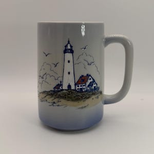 May include: A tall ceramic mug with a light blue gradient base. The front features a detailed blue ink illustration of a lighthouse and coastal buildings on a sandy dune, accented with small orange roof details and flying birds.