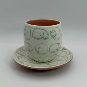 May include: A ceramic cup and saucer set. The cup and saucer are off-white with a pink rim. The cup and saucer are decorated with blue line drawings of sleeping cats. The cup is cylindrical.