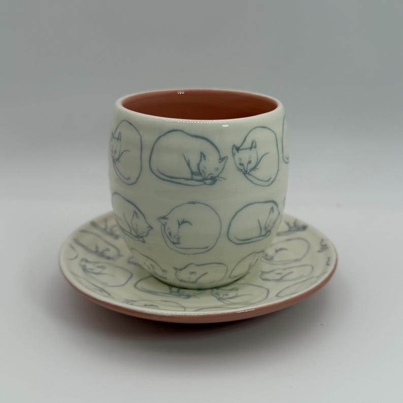May include: A ceramic cup and saucer set. The cup and saucer are off-white with a pink rim. The cup and saucer are decorated with blue line drawings of sleeping cats. The cup is cylindrical.