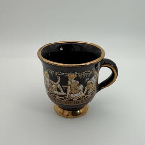 May include: A black ceramic mug featuring a gold-toned rim, handle, and pedestal base. The exterior is decorated with a wrap-around illustration of figures in classical attire, accented with gold, white, and green details and a Greek key border.