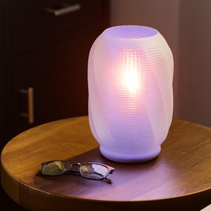 May include: A white, textured lamp with a soft glow, sitting on a wooden table. The lamp has a unique, ribbed design and emits a warm, inviting light. A pair of eyeglasses rests on the table near the lamp. The lamp is a decorative home accent.