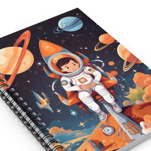 May include: A spiral bound notebook with a colorful illustration of an astronaut in a rocket ship flying through space. The astronaut is wearing a white spacesuit and a blue helmet. The background is a dark blue with stars, planets, and a comet.