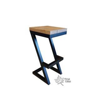 Op de afbeelding: Een zwarte metalen barstoel met een houten zitting. De stoel heeft een Z-vormig frame en een voetensteun. De zitting is gemaakt van natuurlijk hout met een lichtbruine afwerking.