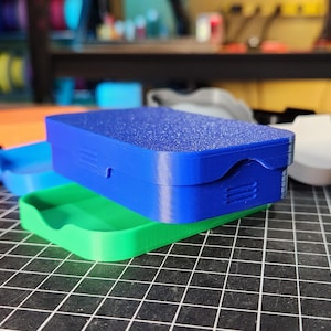 May include: A collection of brightly coloured, 3D-printed boxes in red, orange, blue, and green. The top box is blue with a textured surface. The boxes are on a black grid.