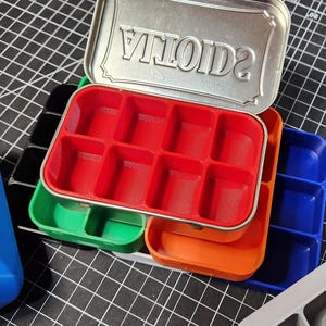May include: An open silver tin with the word "VITOIDS" embossed on the lid, revealing a red compartmented tray. Several other colorful trays in green, orange, and blue are visible, suggesting a storage or organizational product.