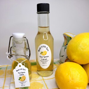 May include: Two bottles of pure lemon extract, one small and one large, with labels that say "Made with Love, Pure Lemon Extract". The bottles are sitting on a white and yellow checkered tablecloth with lemons.