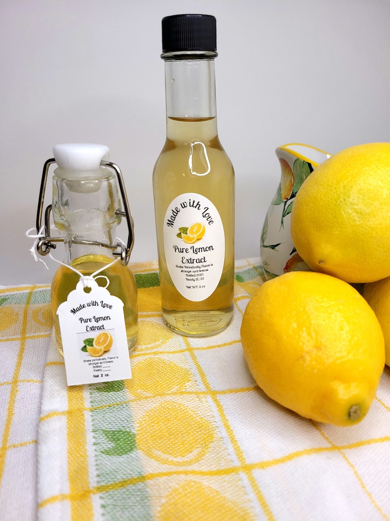May include: Two bottles of pure lemon extract, one small and one large, with labels that say "Made with Love, Pure Lemon Extract". The bottles are sitting on a white and yellow checkered tablecloth with lemons.