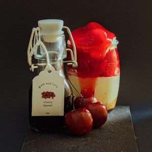 May include: A glass bottle of cherry extract with a white tag that reads "Made with Love" and "Cherry Extract 2oz." Next to it is a glass jar with a layered dessert topped with red cherry sauce. Two fresh cherries sit in front.