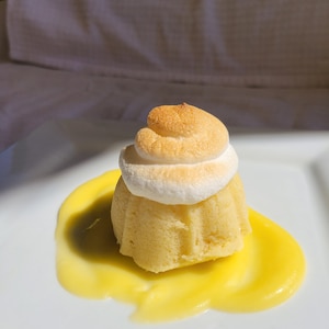 May include: A small, round cake topped with a swirl of white meringue, served on a white plate with a pool of yellow lemon curd.