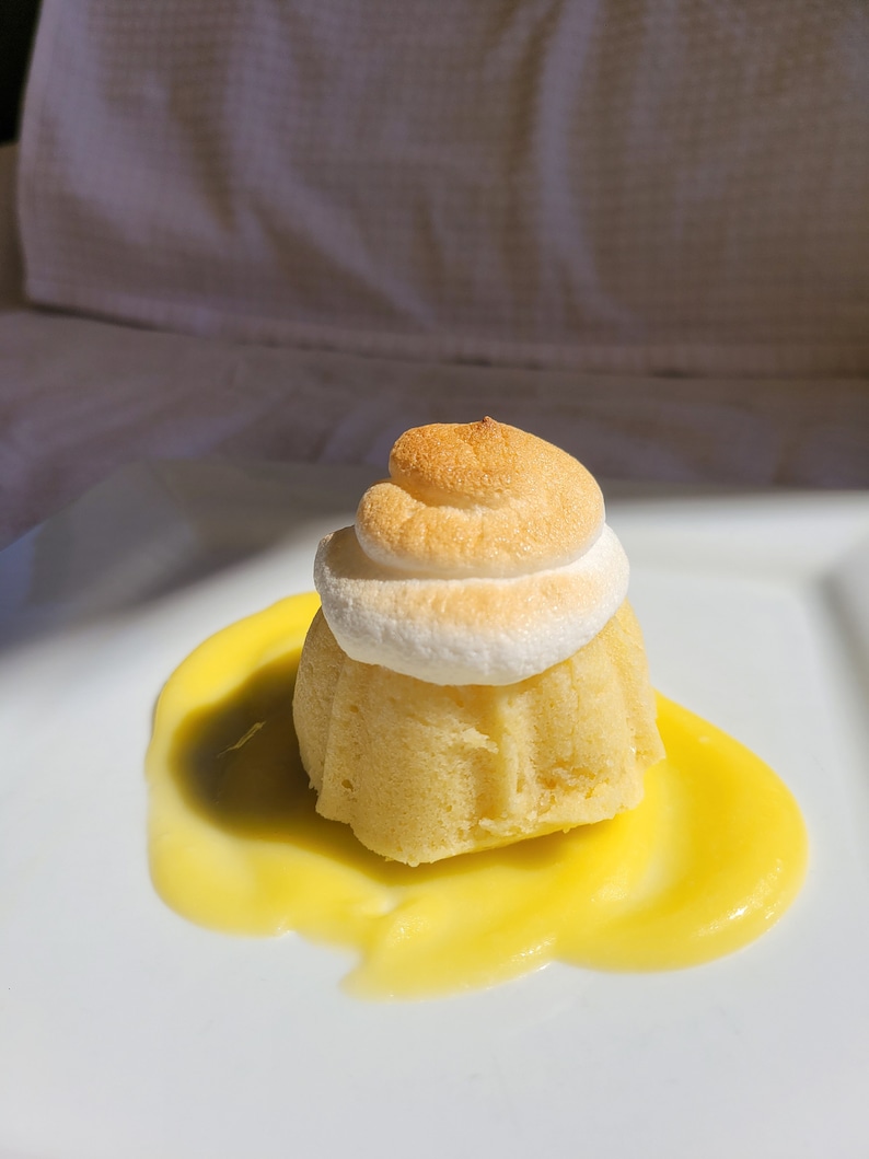 May include: A small, round cake topped with a swirl of white meringue, served on a white plate with a pool of yellow lemon curd.