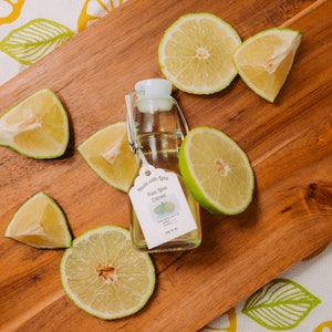 May include: A clear glass bottle with a white label that reads "Made with Love Pure Lime Extract 2 oz". The bottle is sitting on a wooden cutting board with lime slices.
