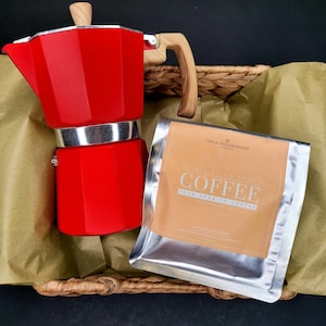 Puede incluir: Una cafetera italiana roja con asa y parte superior de madera, junto a un paquete de granos de café. La cafetera está en una cesta tejida con papel marrón. El paquete de café tiene la palabra "COFFEE".